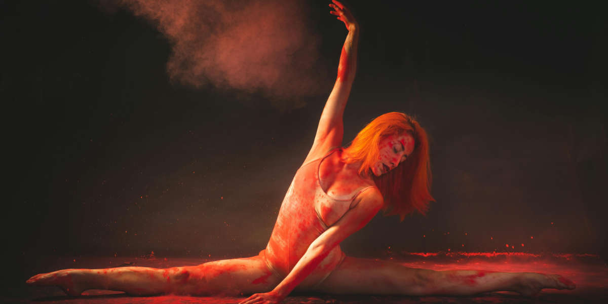 Eine Frau sitzt im Spagat auf den Boden. Auf ihr und um sie herum ist rotes Pulver.