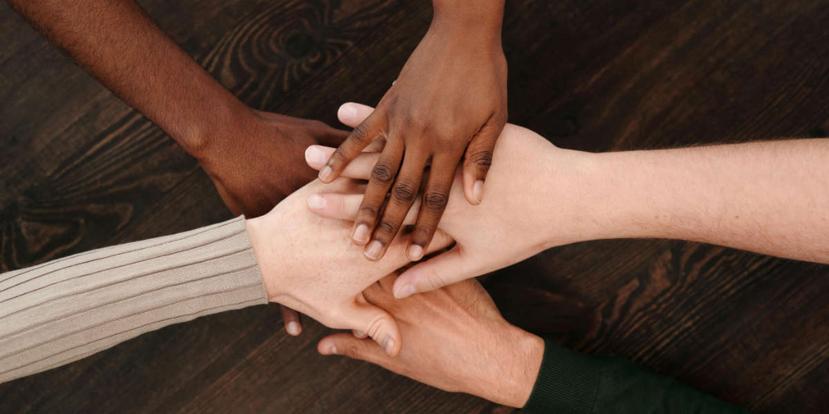 Fünf Hände liegen aufeinander auf einem Tisch. Die Hände haben unterschiedliche Hautfarben.