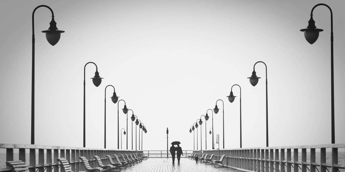 Ein Holzsteg mit Bänken und Laternen . In der Ferne gehen zwei Personen unter einem Regenschirm. 