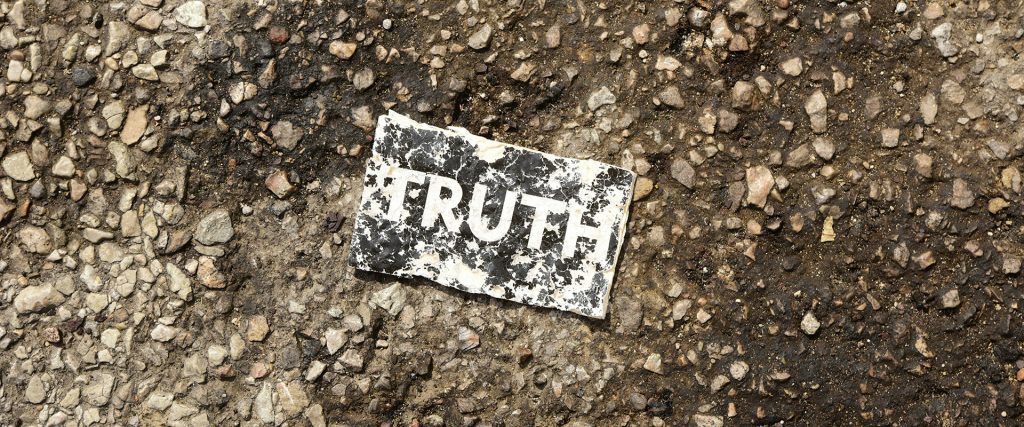 Ein zerknittertes und abgenutztes Stück Papier mit dem Wort „TRUTH“ liegt auf einem groben, steinigen Boden.