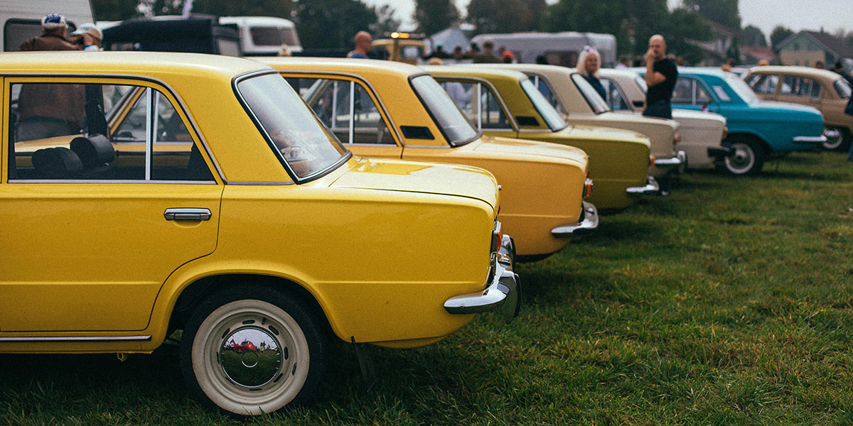 Mehrere alte Kleinwagen stehen in einer Reihe auf einer Wiese. Die Fahrzeuge sind in verschiedenen Farben lackiert, darunter Gelb, Beige und Blau. Im Hintergrund stehen einige Menschen zwischen Wohnwagen und weiteren Autos. Hinter ihnen sind Bäume und Gebäude zu sehen.