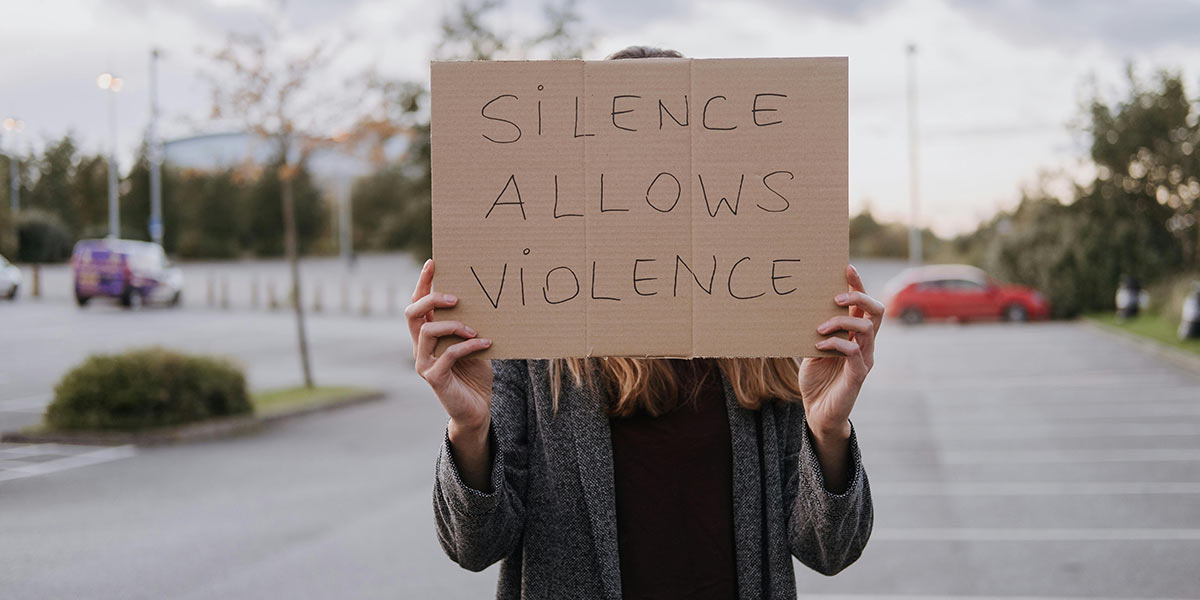 Eine Person hält ein Pappschild vor ihr Gesicht, auf dem steht: „SILENCE ALLOWS VIOLENCE“. Im Hintergrund ist ein Parkplatz mit einigen Autos und Bäumen zu sehen.