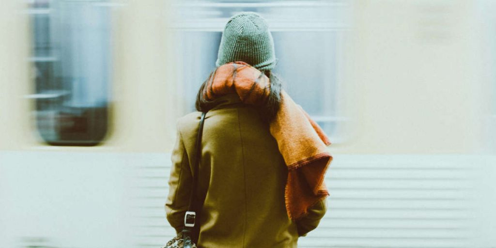 Rückansicht einer Person mit Mütze, Schal und Tasche, vor ihr fährt ein Zug vorbei