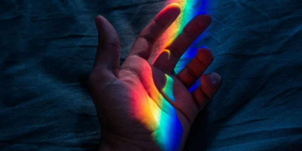 Eine Hand liegt geöffnet in einem dunklen Raum auf einem Tuch. Auf die Hand fällt ein Lichtstreifen in Regenbogenfarben